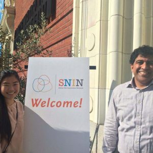 two students standing next to a sign reading "Safety Net Innovation Network: Welcome!"