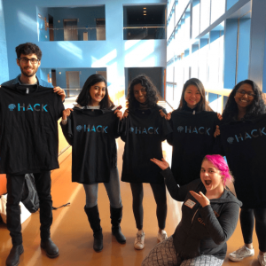 Five people hold up black shirts that say "Hack" while one person with pink hair gestures to them.
