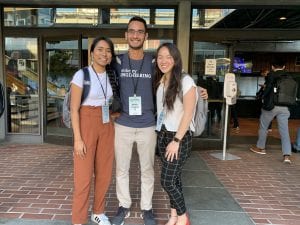 Two young women and a young man with dark hair posed in front of a auditorium hall.